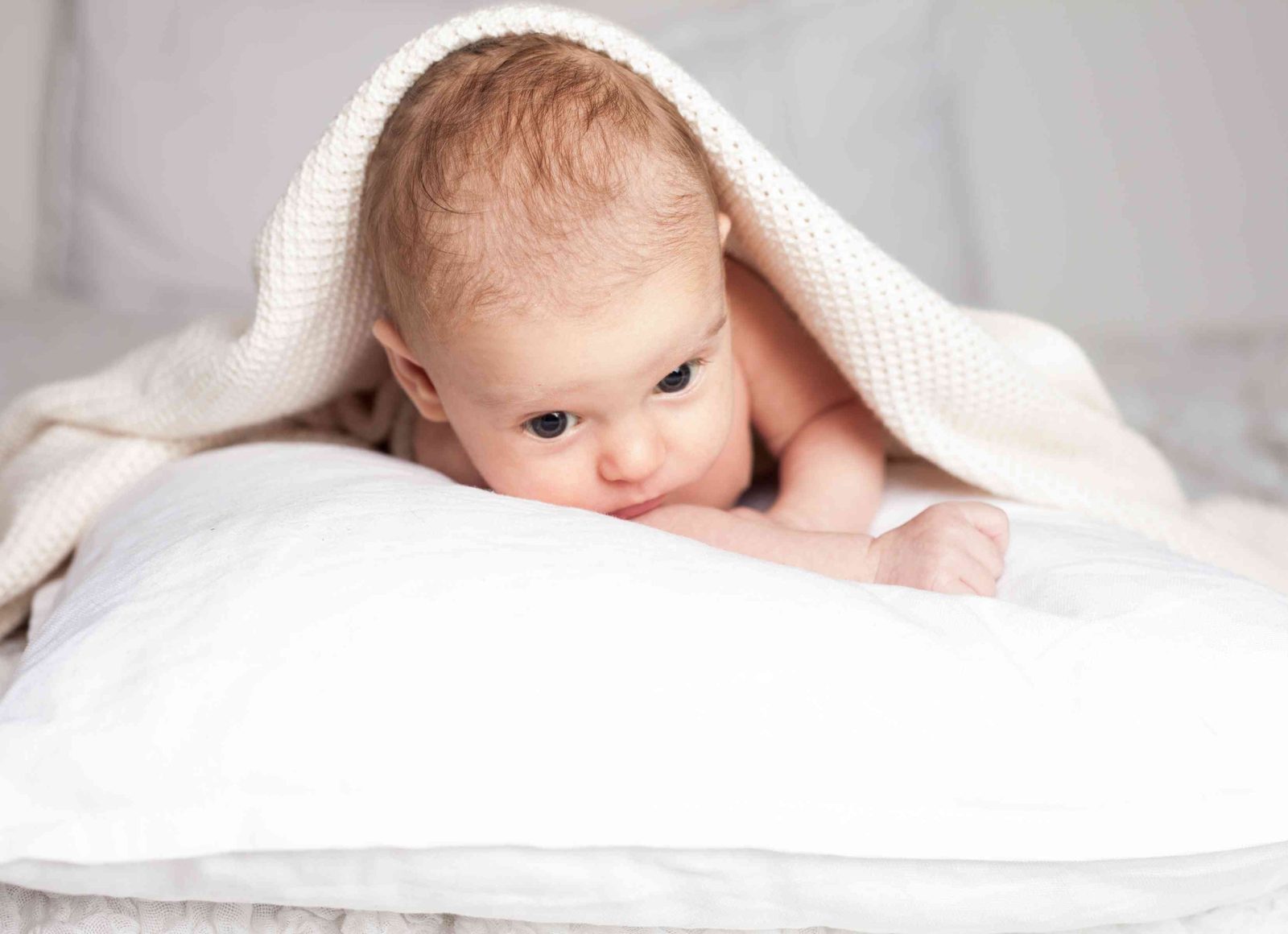 Baby liegt unter Strickdecke auf weißem Kissen und schaut in die Kamera