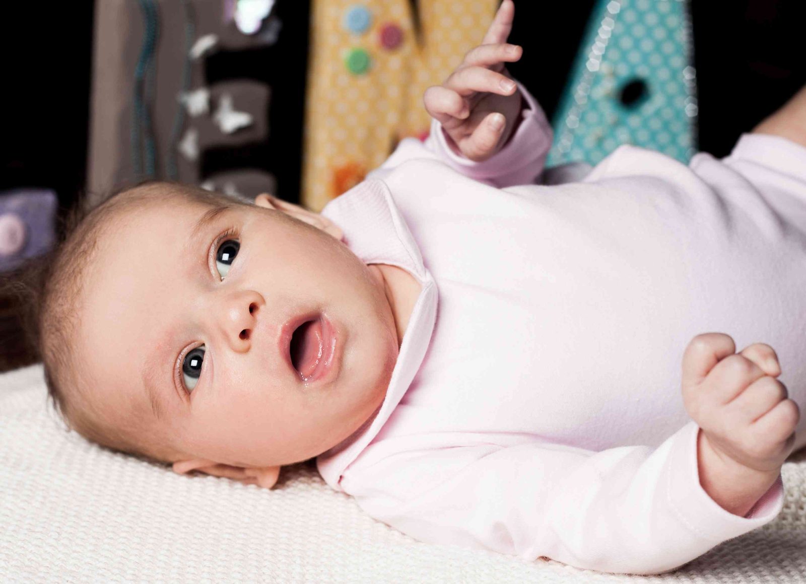 baby in pink onesie lies on a blanket, looking up with an open mouth