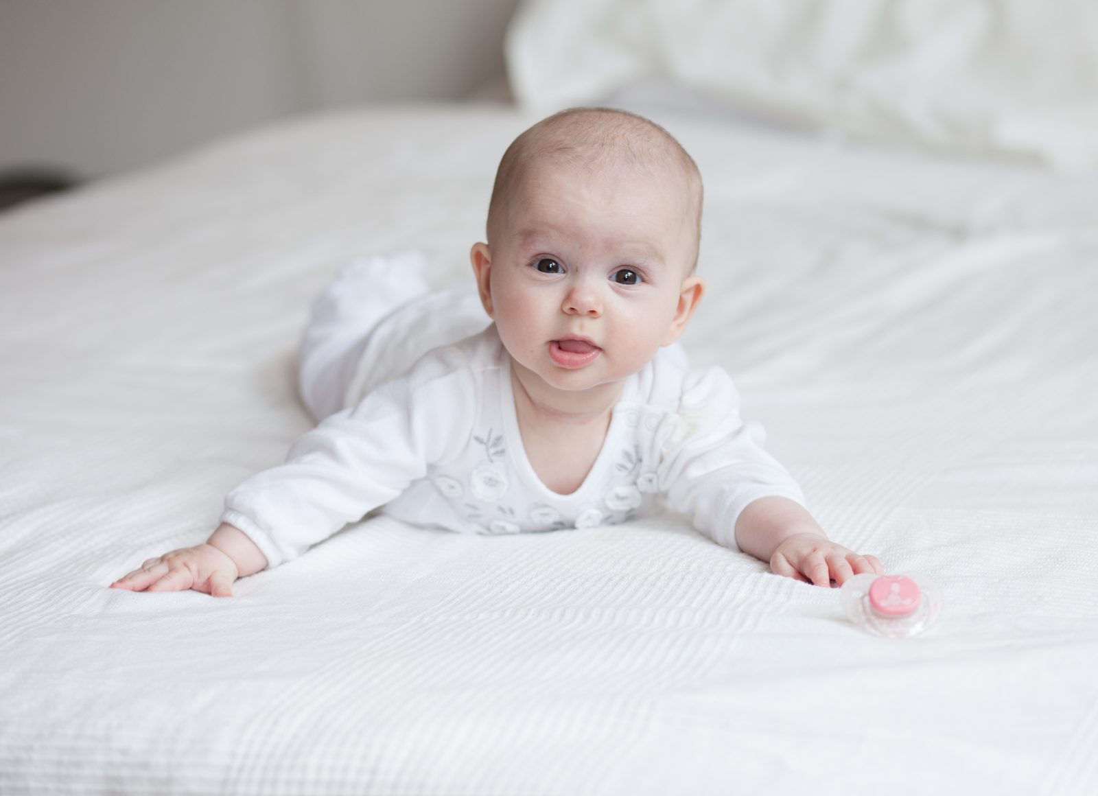 Baby in weißer Kleidung liegt auf weißem Bett und schaut in die Kamera
