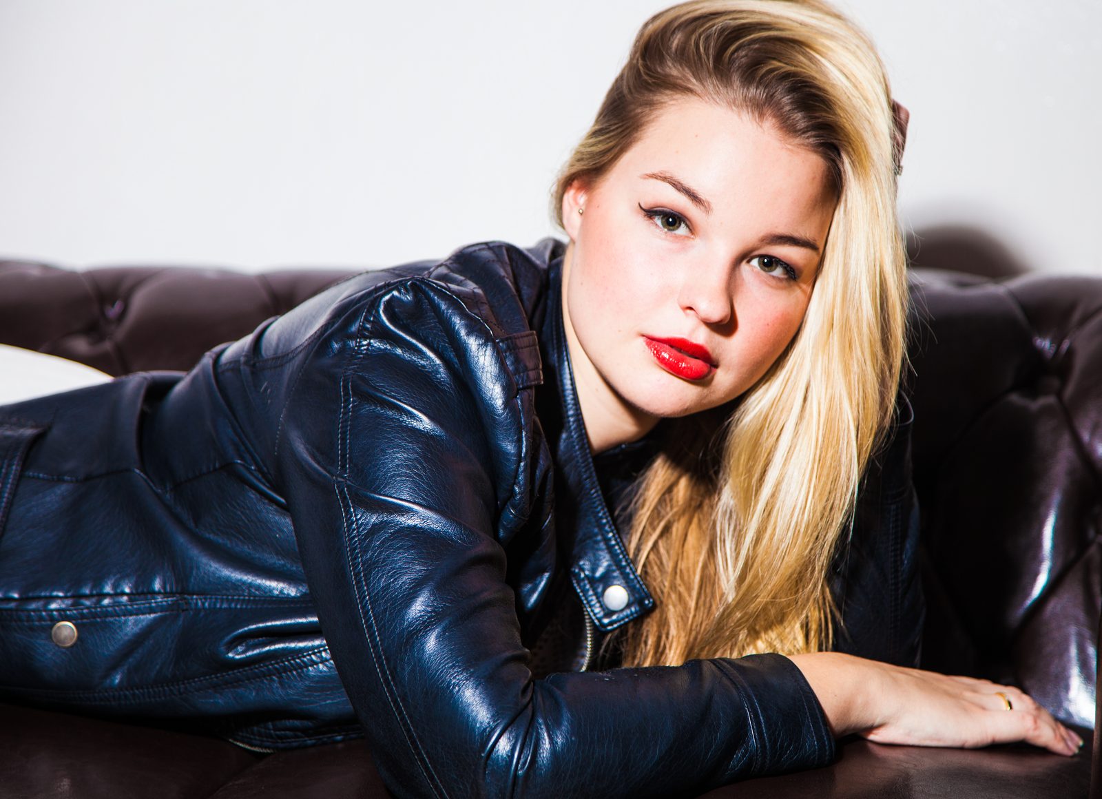 Blonde woman in a leather jacket reclines on a leather sofa, gazing at camera
