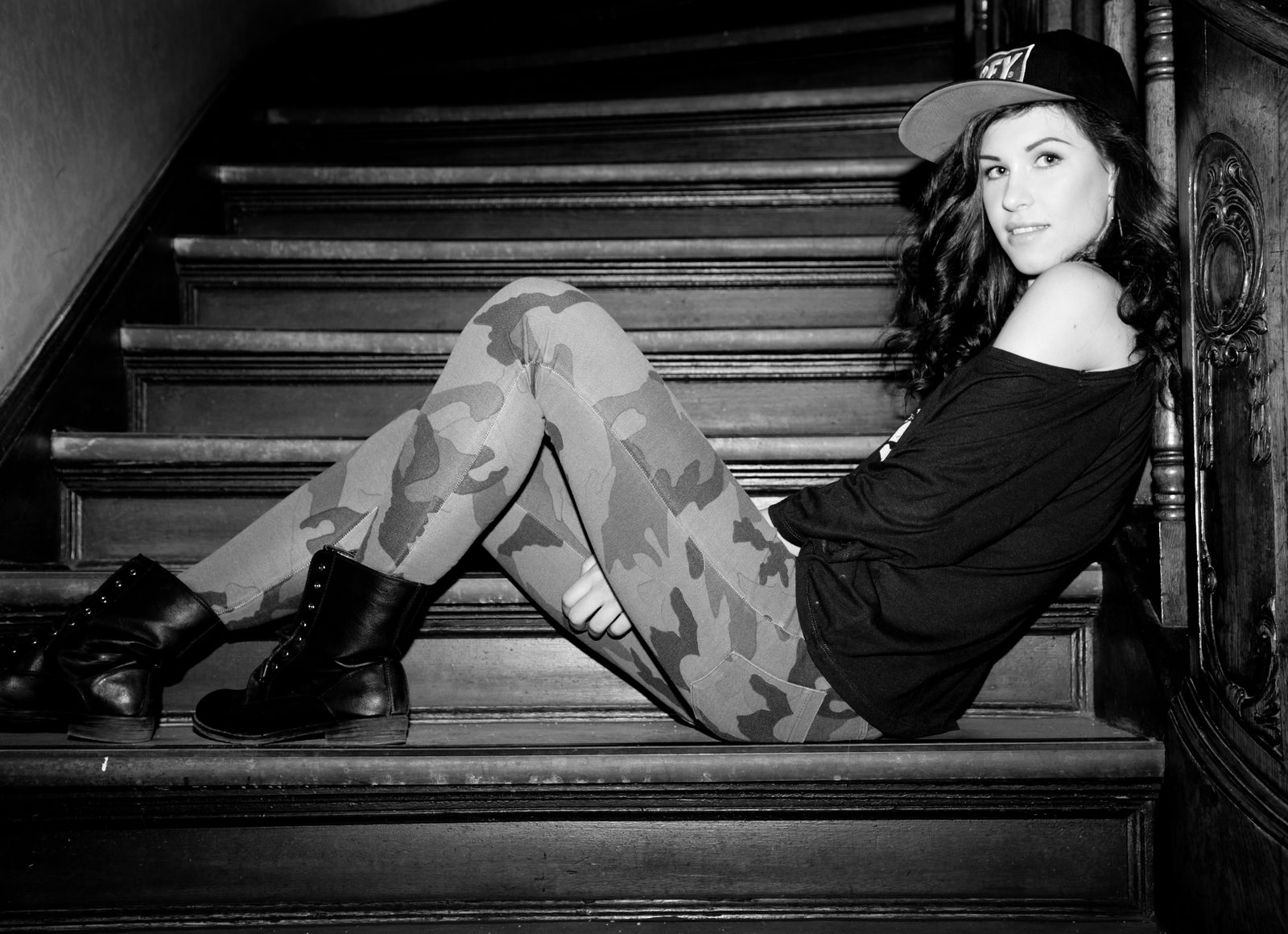 Frau in Cap und Camouflage-Leggings liegt lächelnd auf einer Holztreppe – Schwarz-Weiß-Foto
