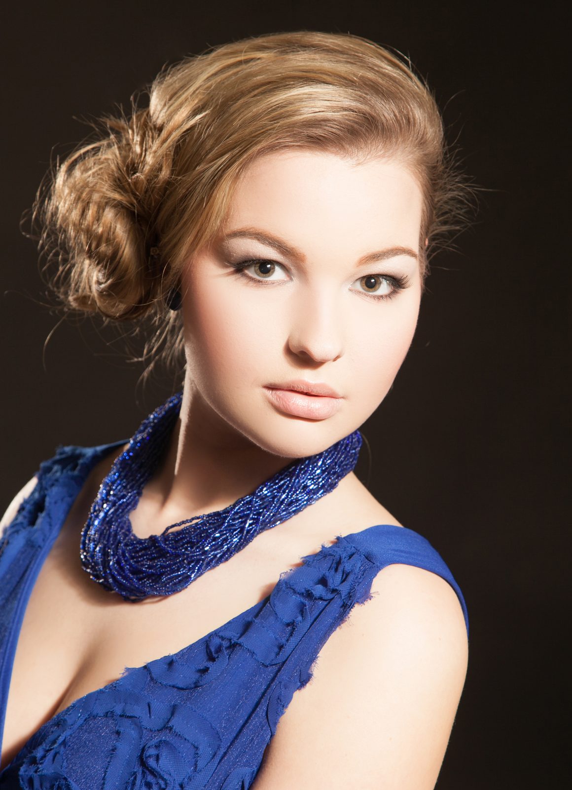 Beauty portrait of a woman in a blue dress with a beaded necklace against a dark backdrop