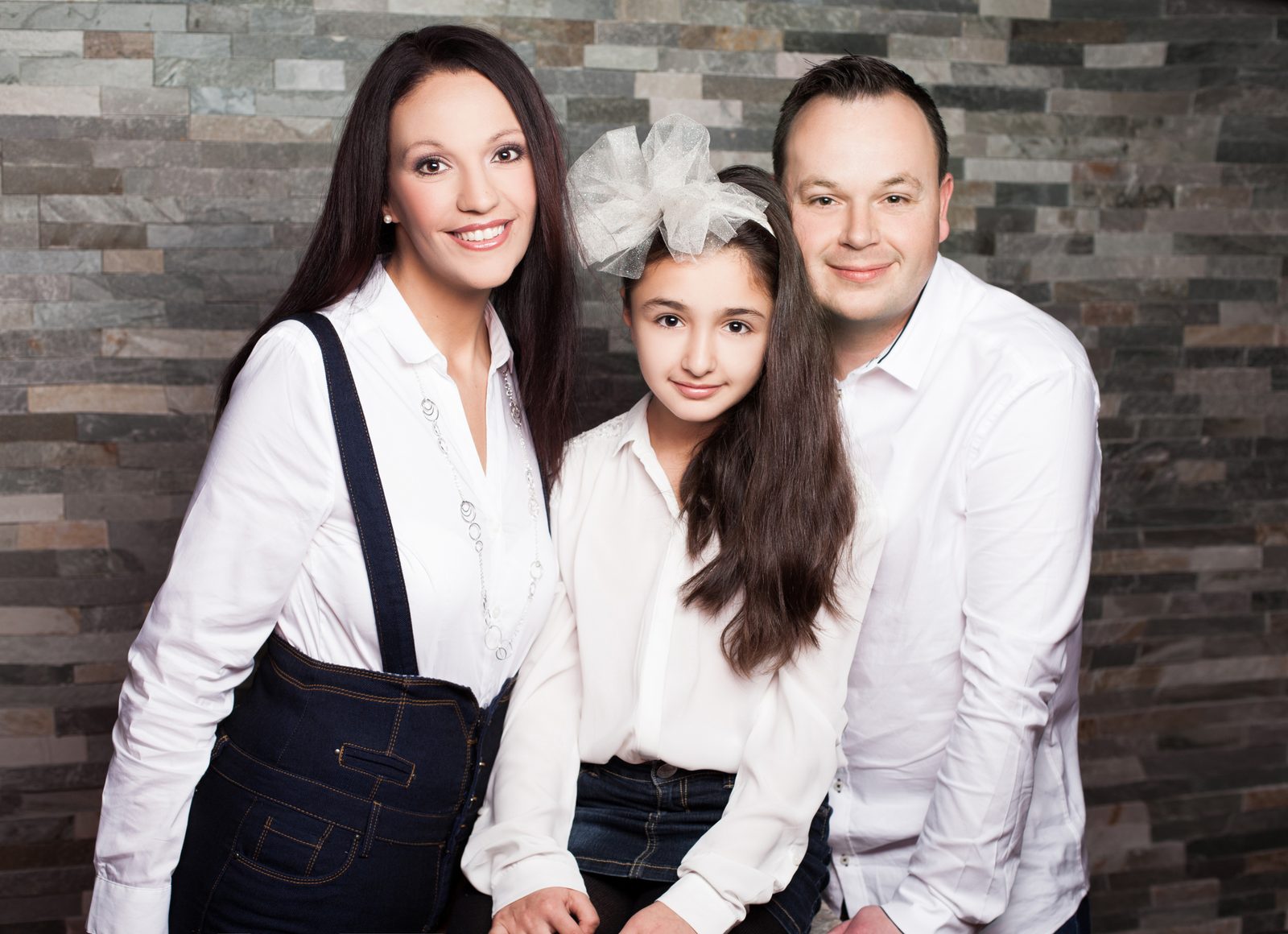 Smiling family of mother, father, and daughter in white outfits against a stone wall