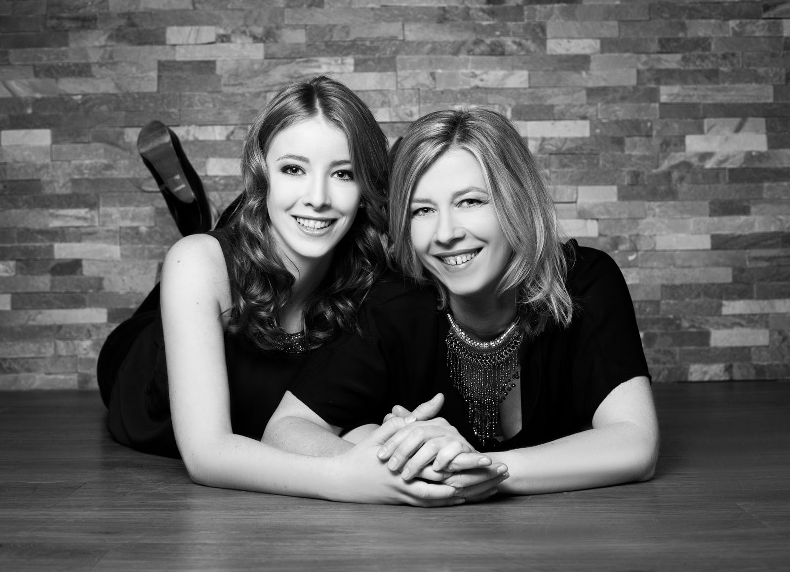 Two smiling women lying side by side holding hands against a stone wall backdrop
