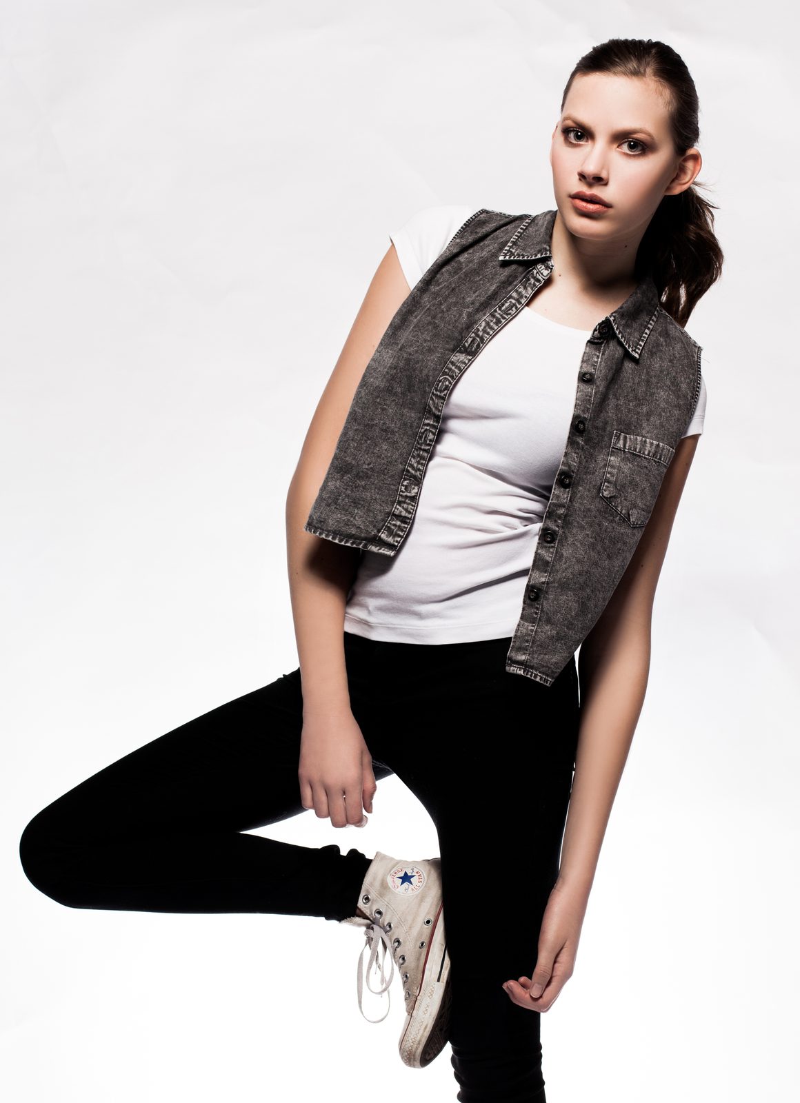 Young woman in denim vest and sneakers posing against a white studio backdrop