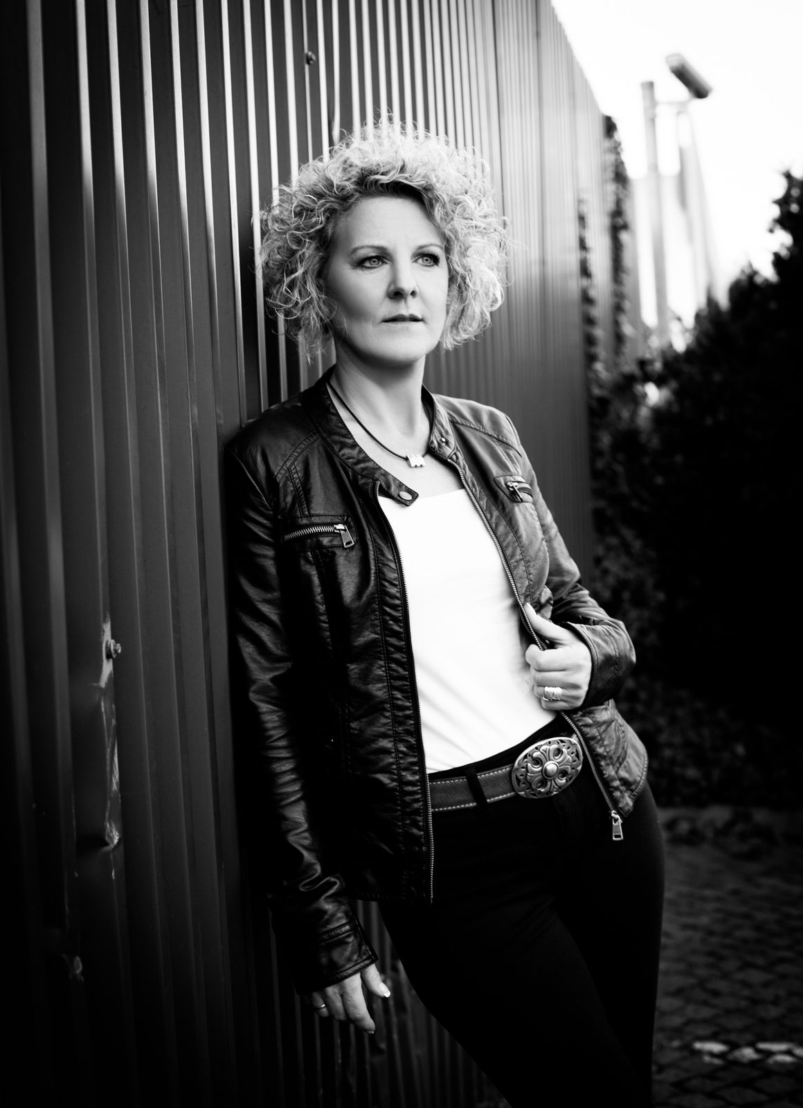 Curly-haired woman in leather jacket leans against corrugated wall—black and white portrait