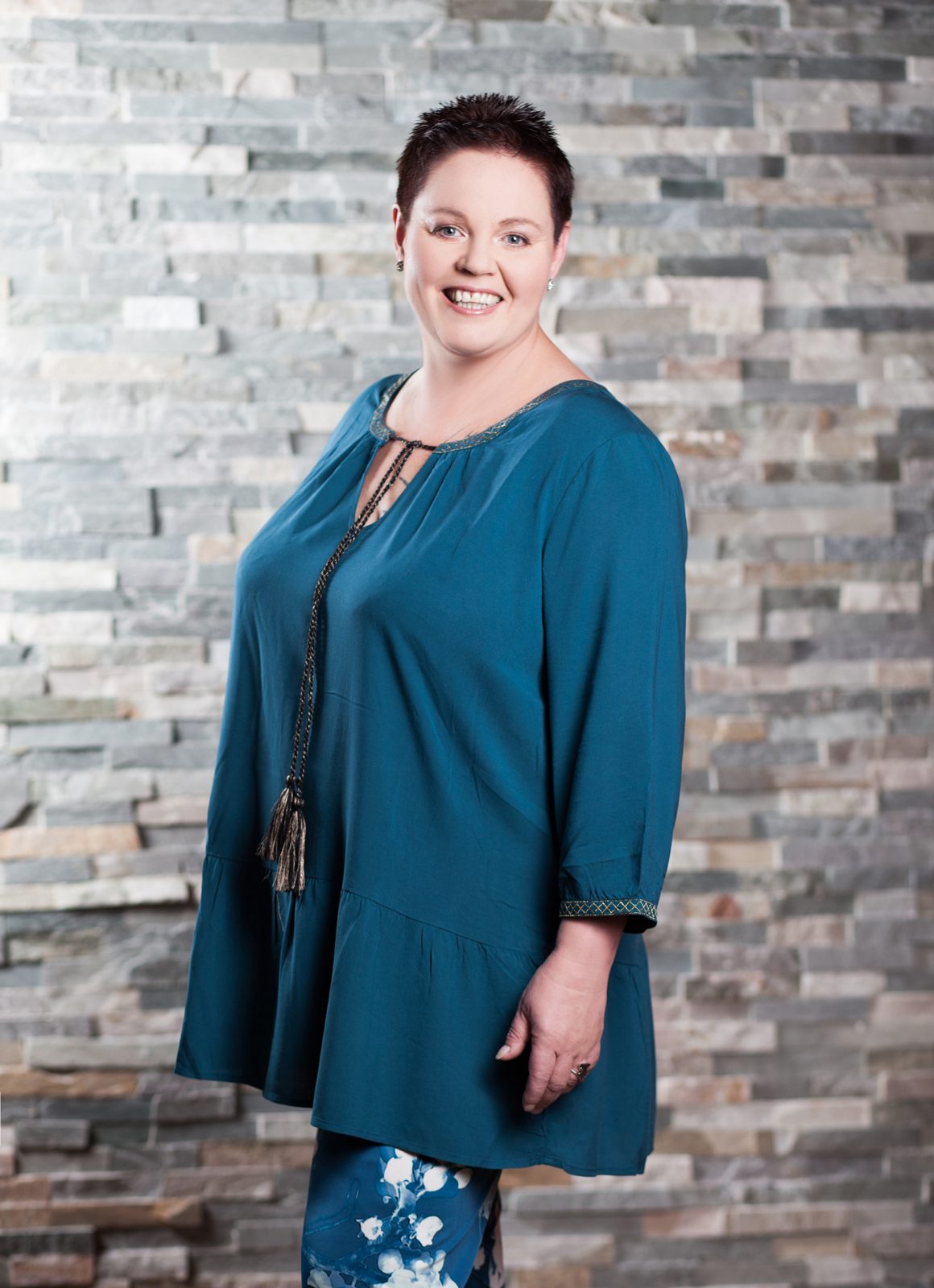 Smiling woman in a teal tunic poses against a stone wall backdrop