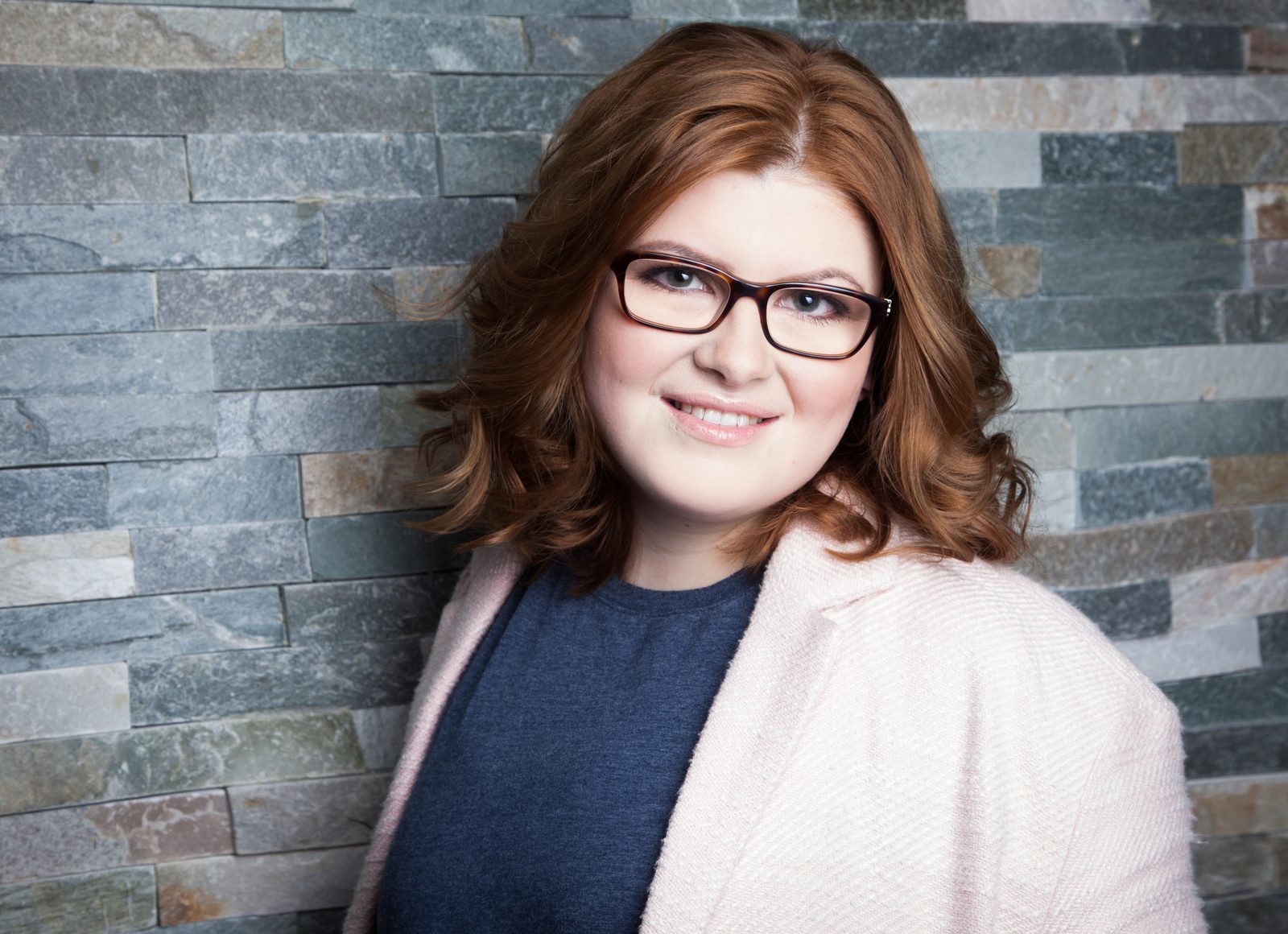 Smiling woman with glasses and pink blazer against a stone wall