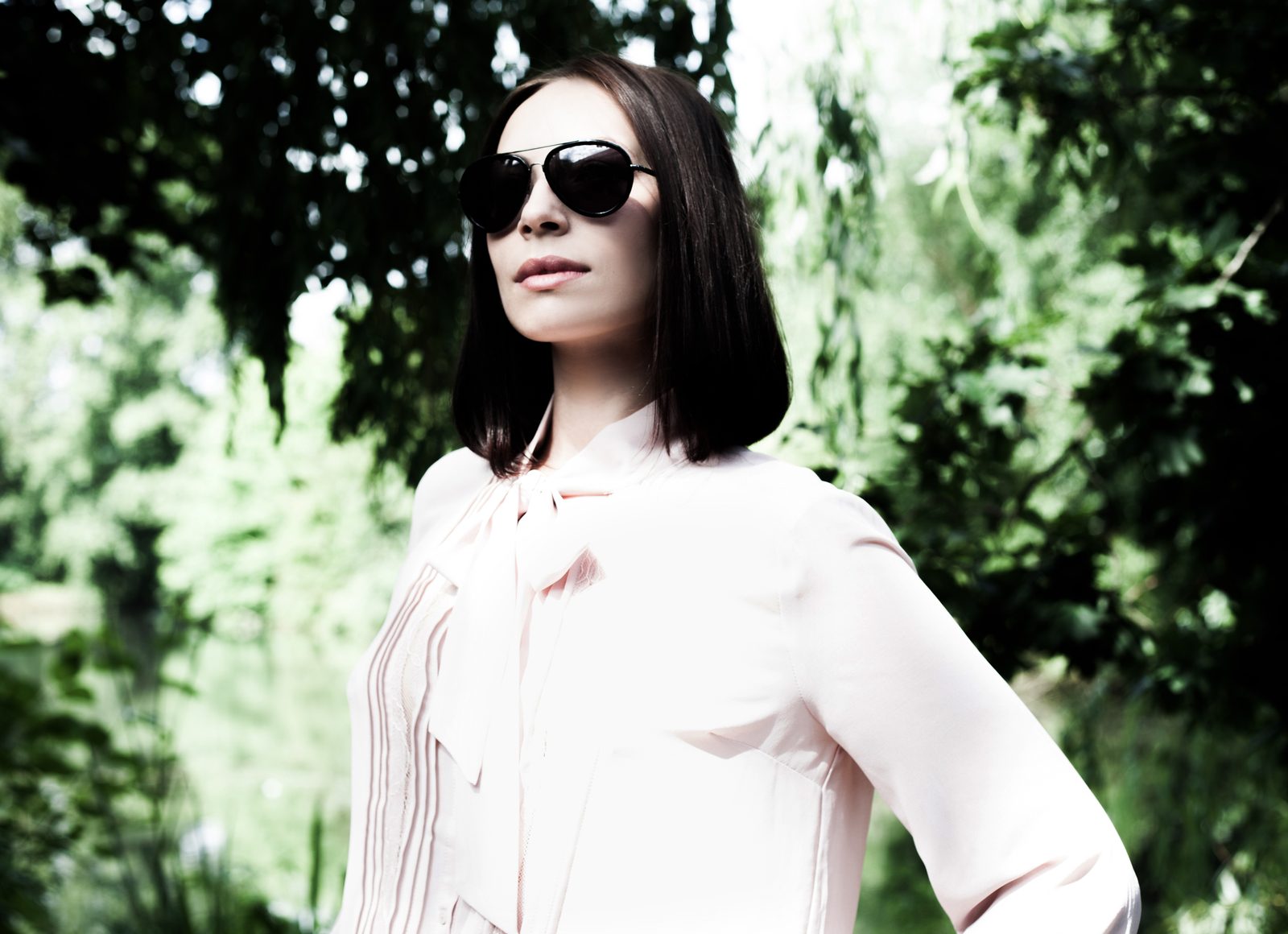 Woman in sunglasses and pale pink blouse posing outdoors against greenery