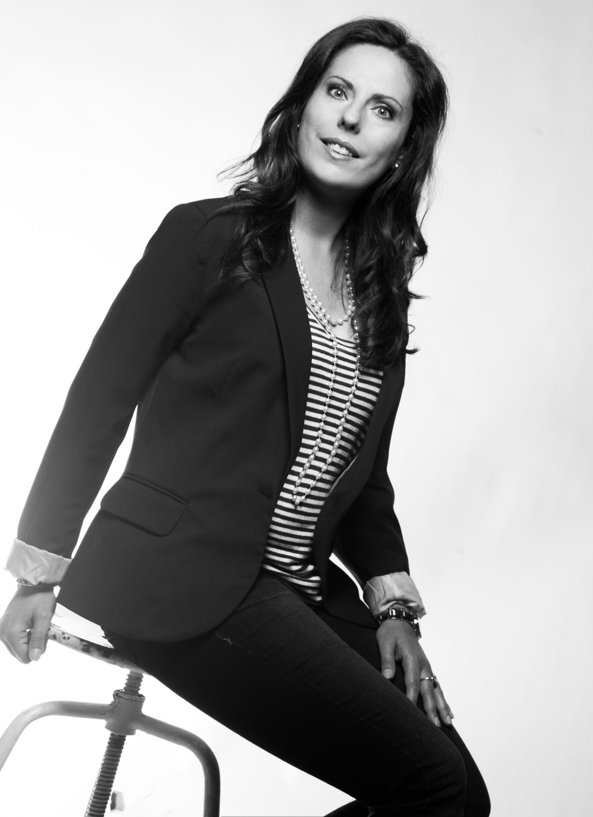 Woman in a black blazer sits smiling on a stool—black-and-white portrait
