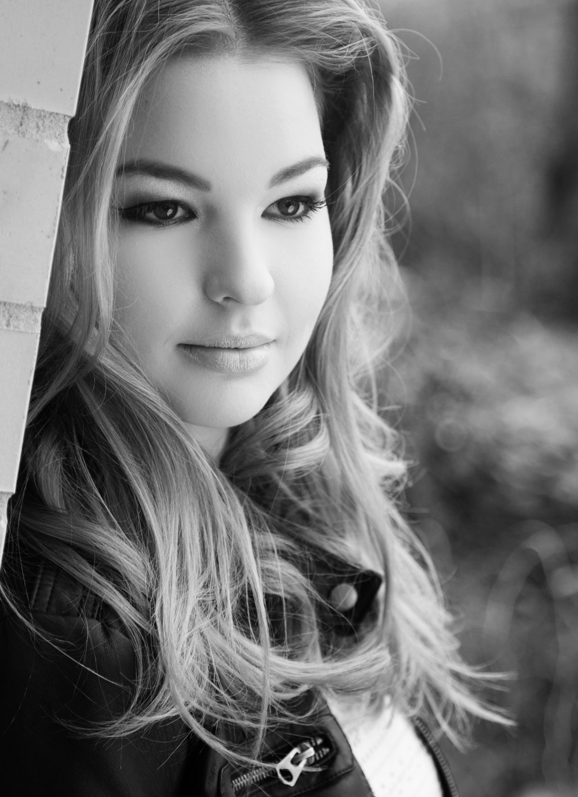 Black-and-white portrait of a young woman with wavy hair leaning against a wall