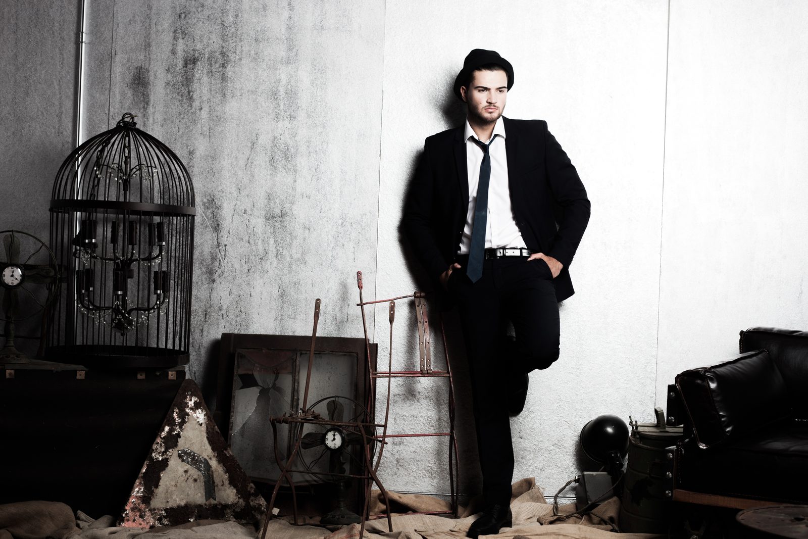Man in a dark suit and hat leaning against a wall amid vintage studio props