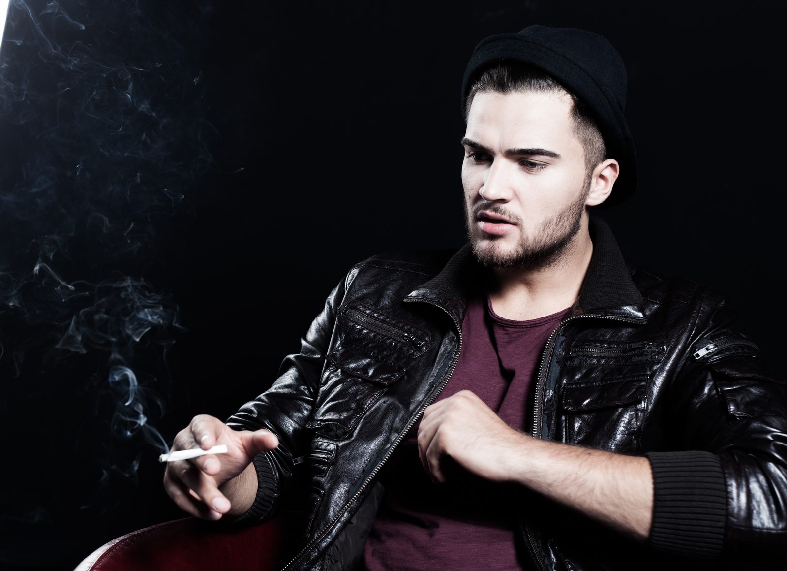 Man in leather jacket and beanie holding a cigarette against dark studio backdrop