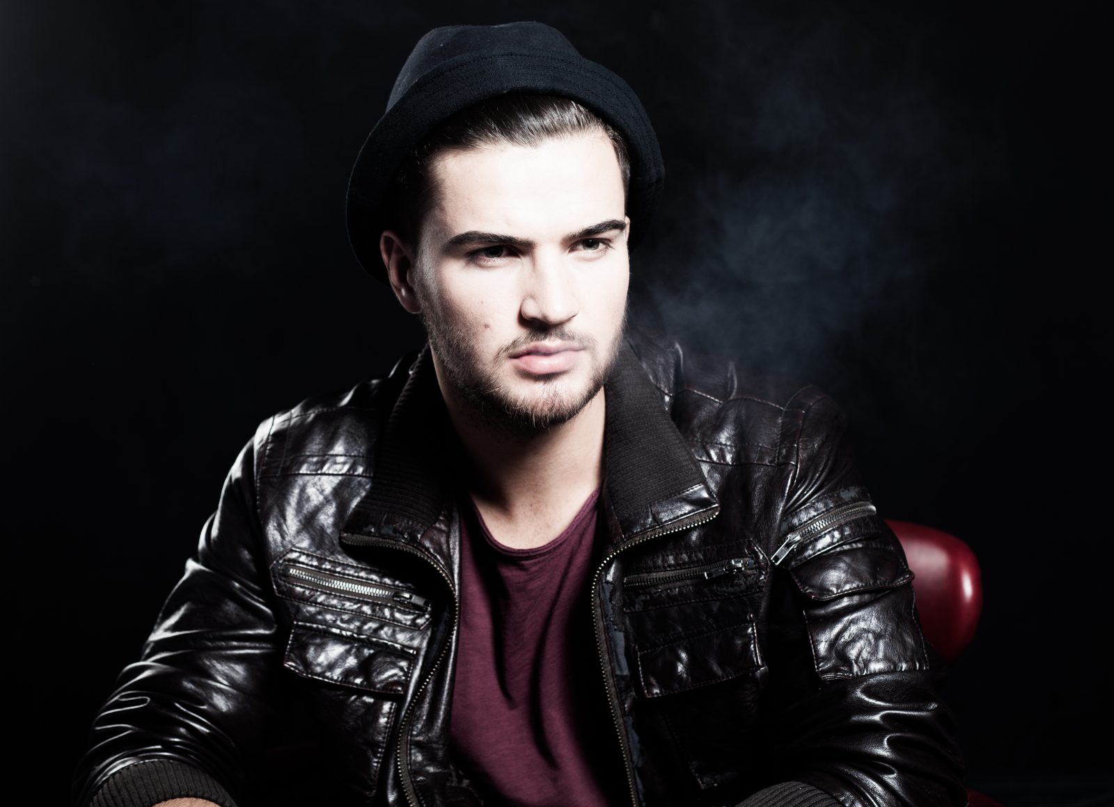 Man in hat and leather jacket against dark studio backdrop, confident gaze