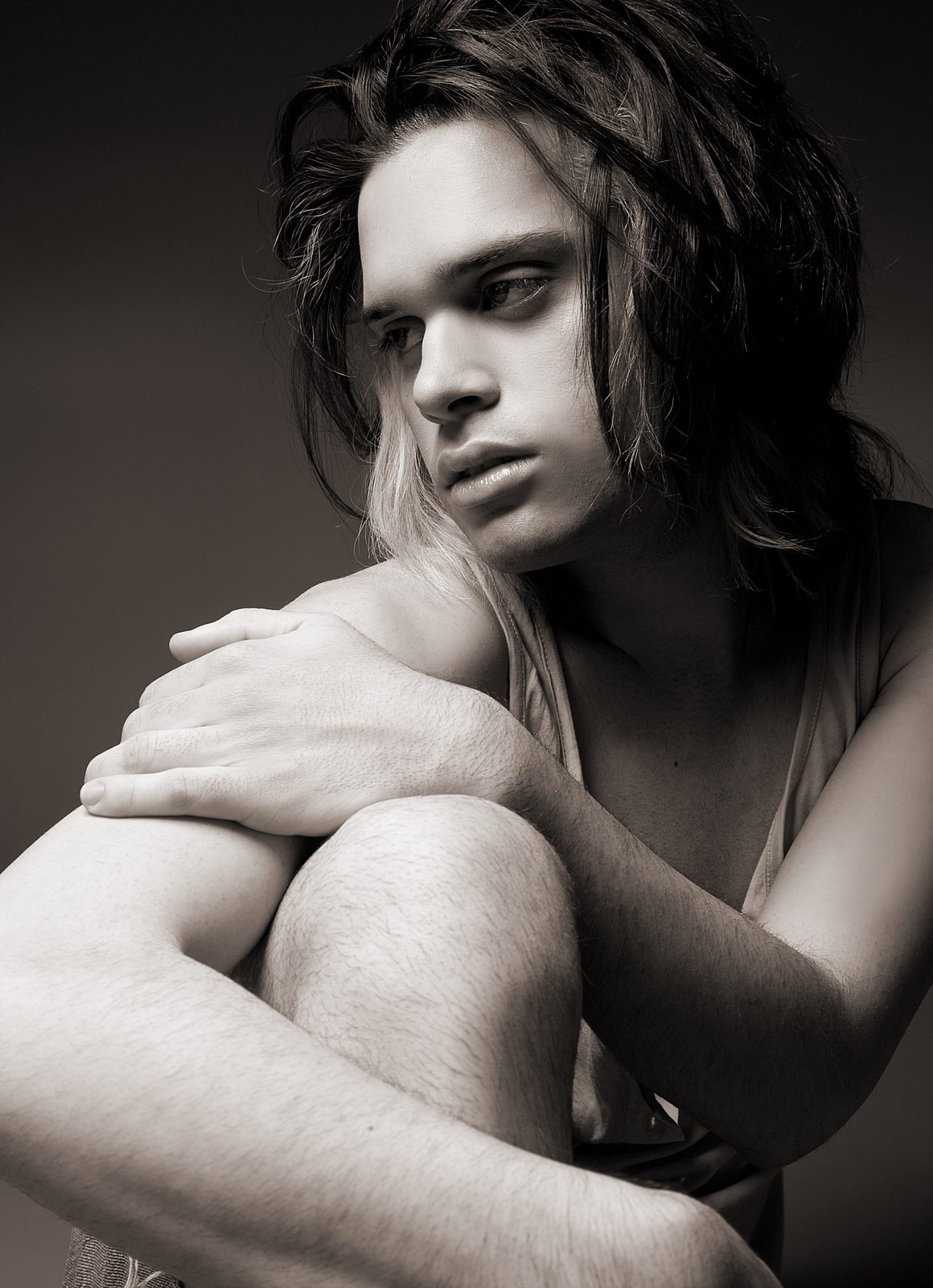 Young man with long hair hugging his knee—black-and-white studio portrait