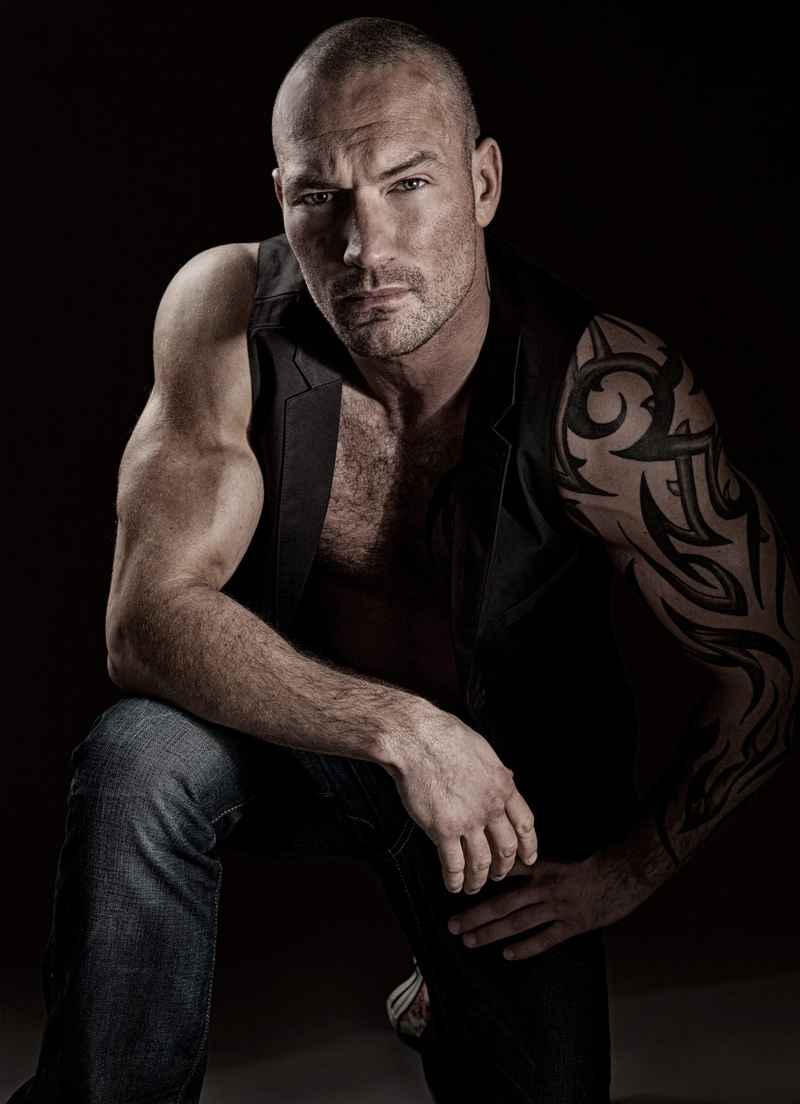 Man with tattoo and vest sitting against a dark studio background